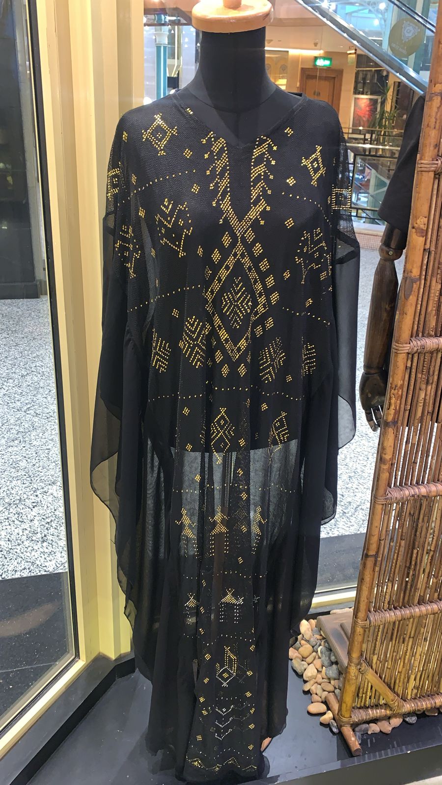 A black abaya with embroidered details and a decorative belt, displayed on a mannequin with an Egyptian-themed backdrop.