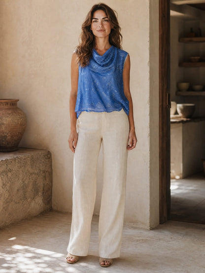 Woman wearing a blue tally sleeveless top and white pants standing in a rustic indoor setting.