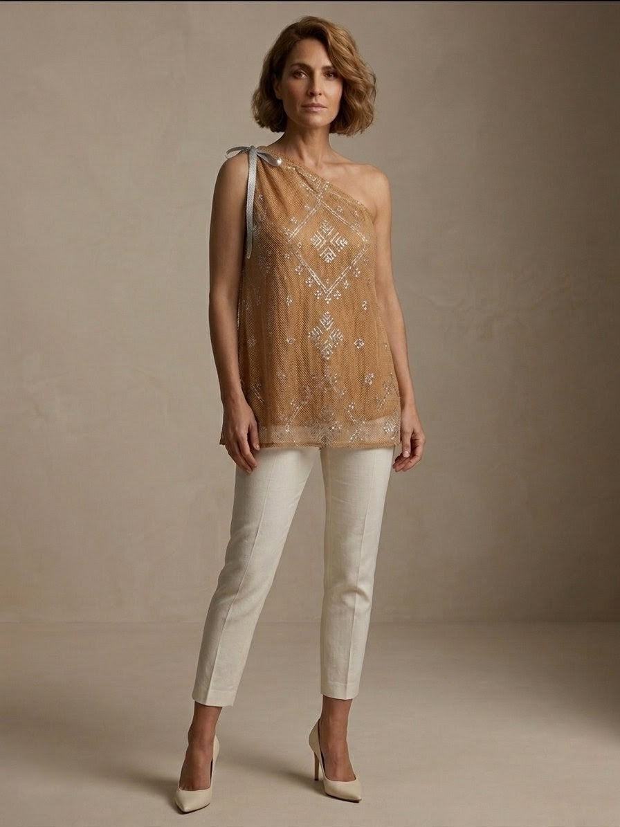 Woman wearing an embroidered tally one-shoulder blouse top and white pants against a neutral background