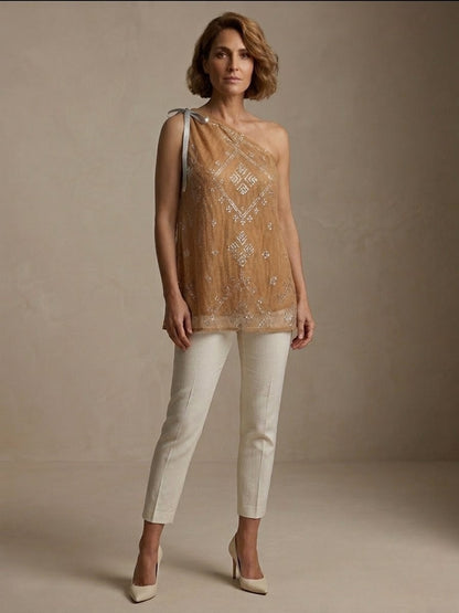 Woman wearing an embroidered tally one-shoulder blouse top and white pants against a neutral background