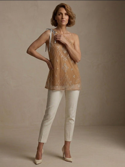 Woman wearing a beige one-shoulder tally top blouse and white pants against a neutral background