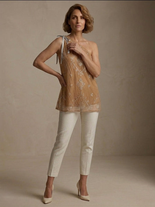 Woman wearing a beige one-shoulder tally top blouse and white pants against a neutral background