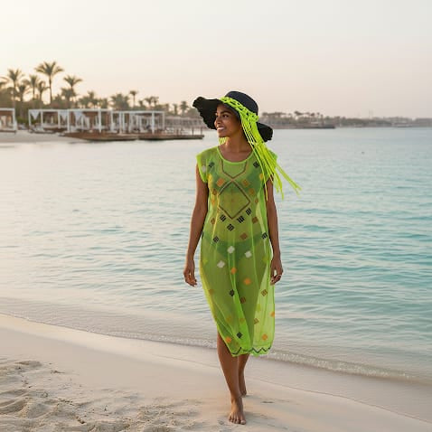 Woman in a green dress and black hat standing on a beach with water and palm trees in the background.