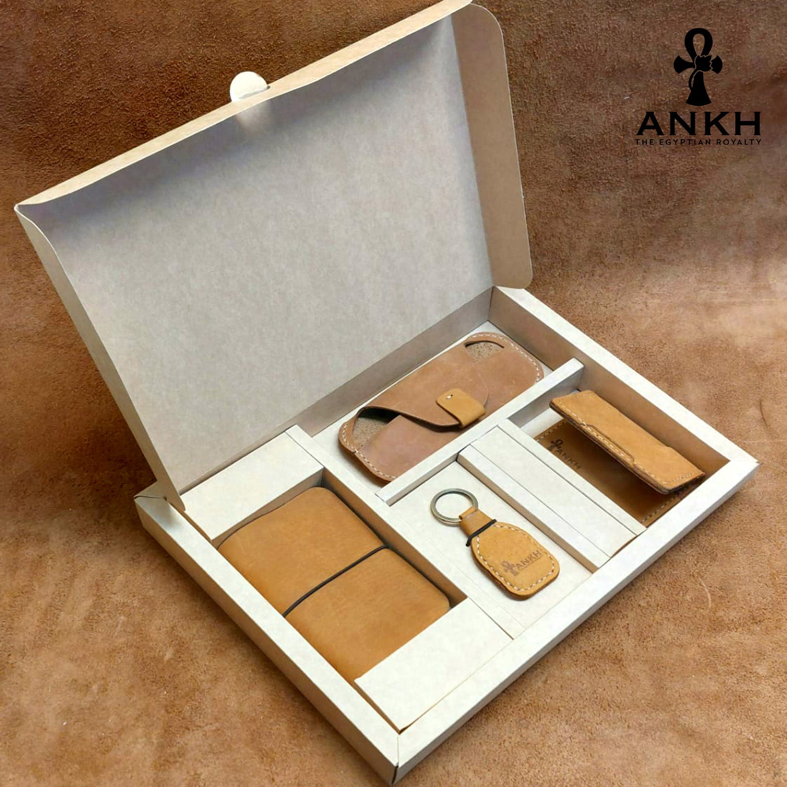 A white box containing four ANKH leather accessories: a passport cover with notebook, a key chain, a glasses cover, and a card holder, displayed on a textured surface. طقم هدية من الجلد الطبيعي الفاخر منقوش برمز العنخ المصري هدية من مصر