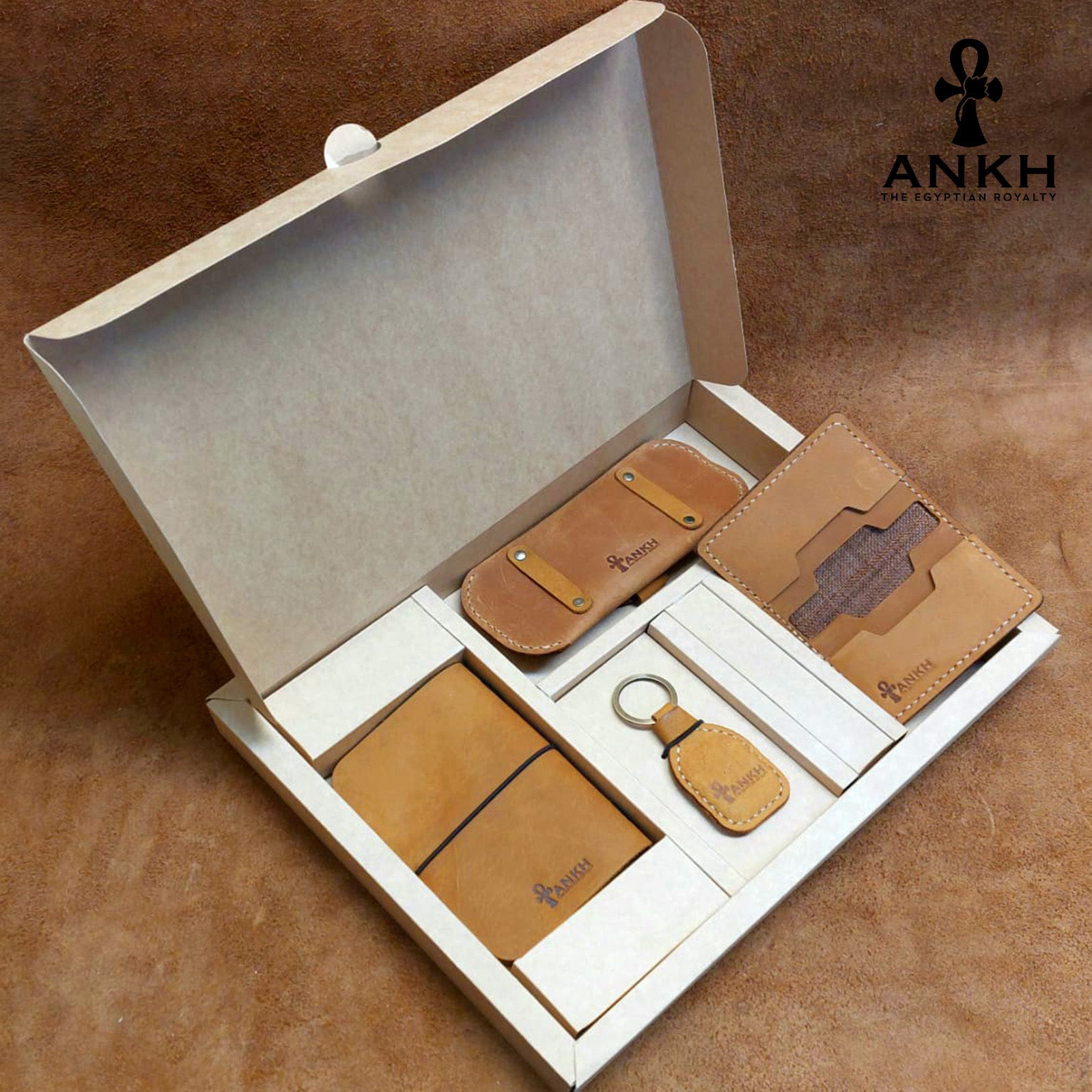 A white box containing four ANKH leather accessories: a passport cover with notebook, a key chain, a glasses cover, and a card holder, displayed on a textured surface. طقم هدية من الجلد الطبيعي الفاخر منقوش برمز العنخ المصري هدية من مصر