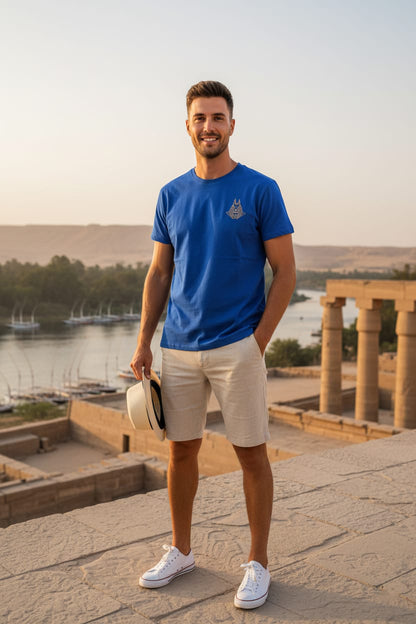 Man in a blue t-shirt and beige shorts standing in front of ancient ruins with a body of water in the background.