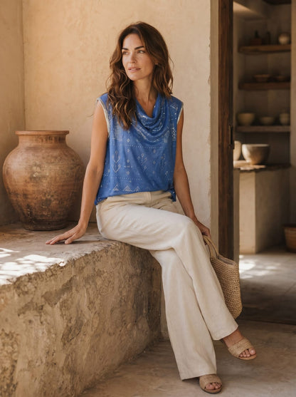 Woman in a blue tally top and beige pants sitting on a stone ledge in a rustic setting.