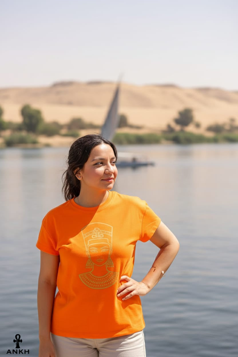 Woman wearing an orange t-shirt with a design, standing by a body of water with a large monument in the background.