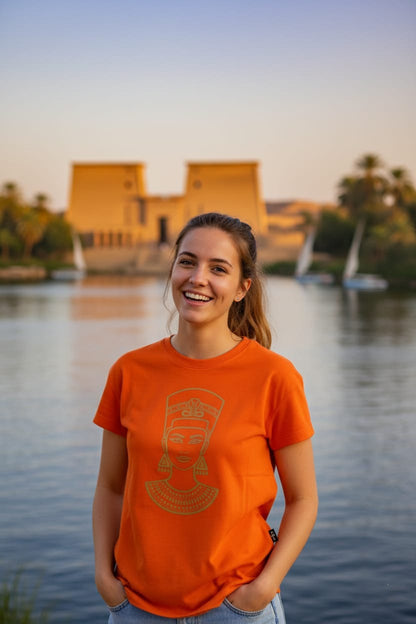 Person wearing an orange t-shirt with a logo, standing by a body of water with a building and palm trees in the background.