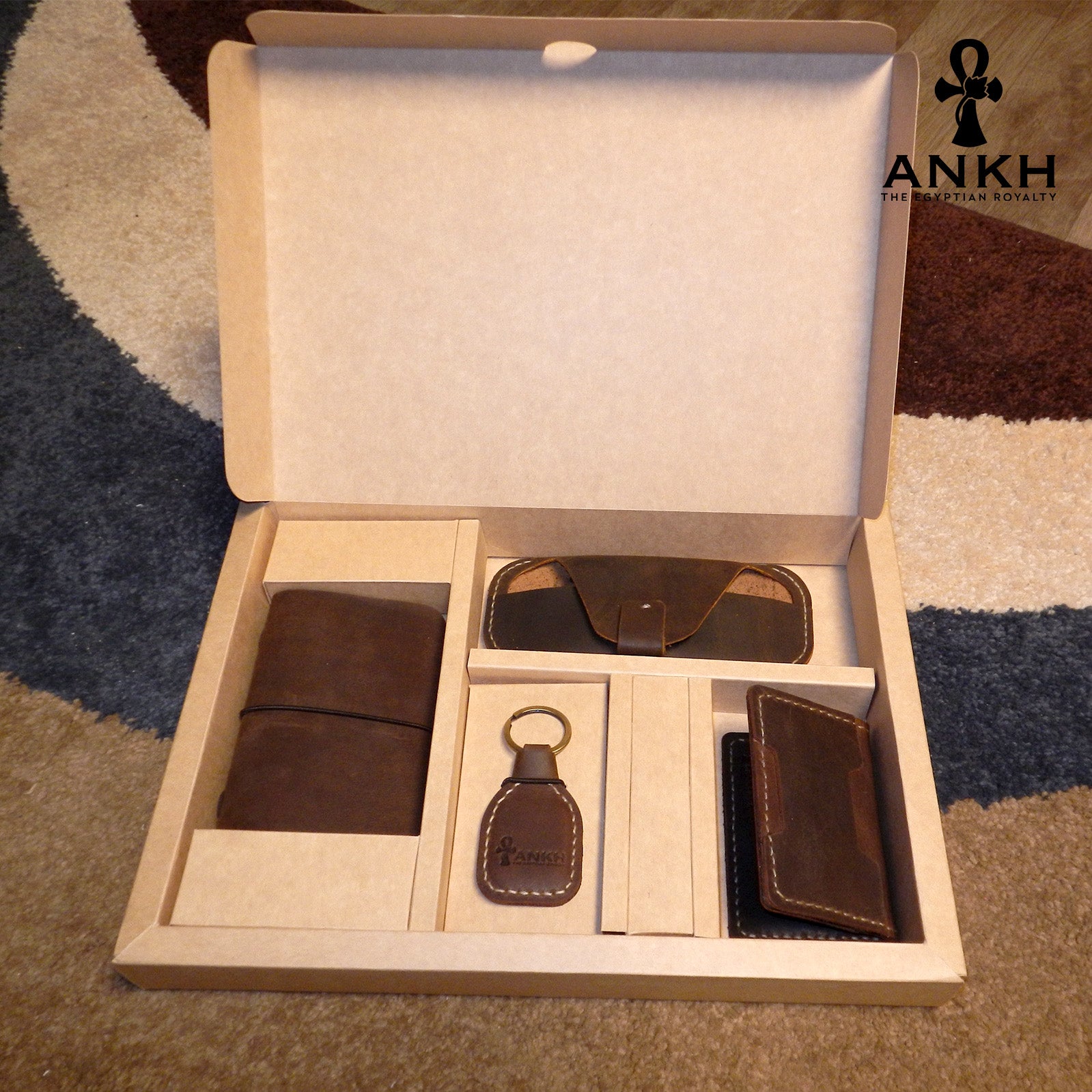 A white box containing four ANKH leather accessories: a passport cover with notebook, a key chain, a glasses cover, and a card holder, displayed on a textured surface. طقم هدية من الجلد الطبيعي الفاخر منقوش برمز العنخ المصري هدية من مصر