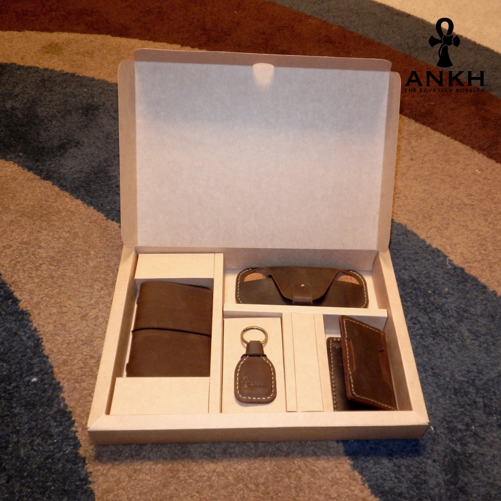 A white box containing four ANKH leather accessories: a passport cover with notebook, a key chain, a glasses cover, and a card holder, displayed on a textured surface. طقم هدية من الجلد الطبيعي الفاخر منقوش برمز العنخ المصري هدية من مصر