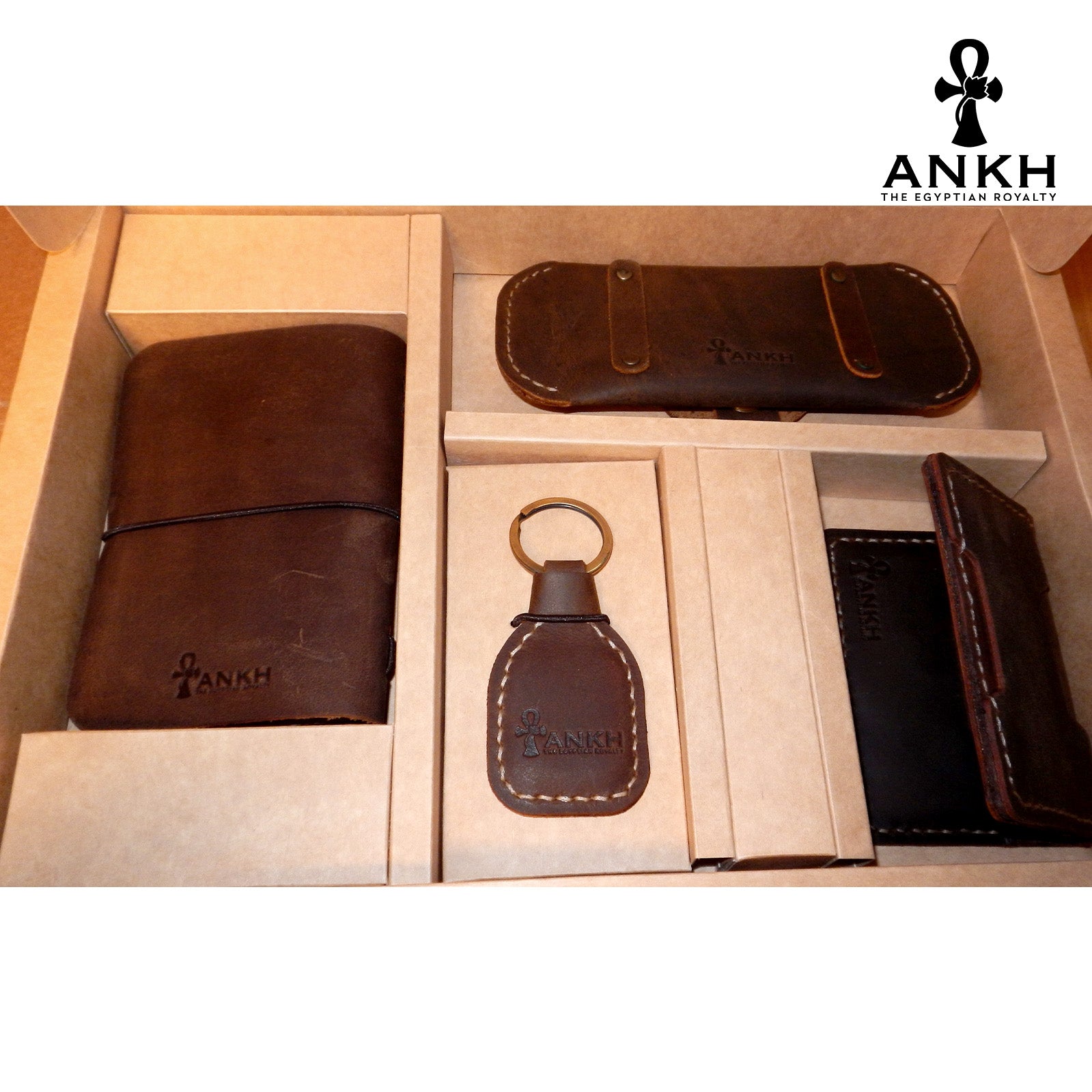 A white box containing four ANKH leather accessories: a passport cover with notebook, a key chain, a glasses cover, and a card holder, displayed on a textured surface. طقم هدية من الجلد الطبيعي الفاخر منقوش برمز العنخ المصري هدية من مصر