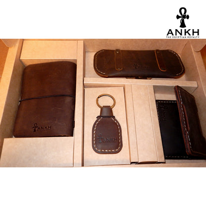 A white box containing four ANKH leather accessories: a passport cover with notebook, a key chain, a glasses cover, and a card holder, displayed on a textured surface. طقم هدية من الجلد الطبيعي الفاخر منقوش برمز العنخ المصري هدية من مصر