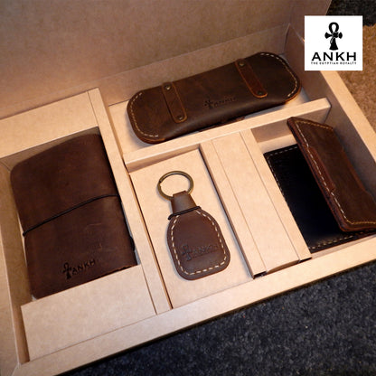 A white box containing four ANKH leather accessories: a passport cover with notebook, a key chain, a glasses cover, and a card holder, displayed on a textured surface. طقم هدية من الجلد الطبيعي الفاخر منقوش برمز العنخ المصري هدية من مصر