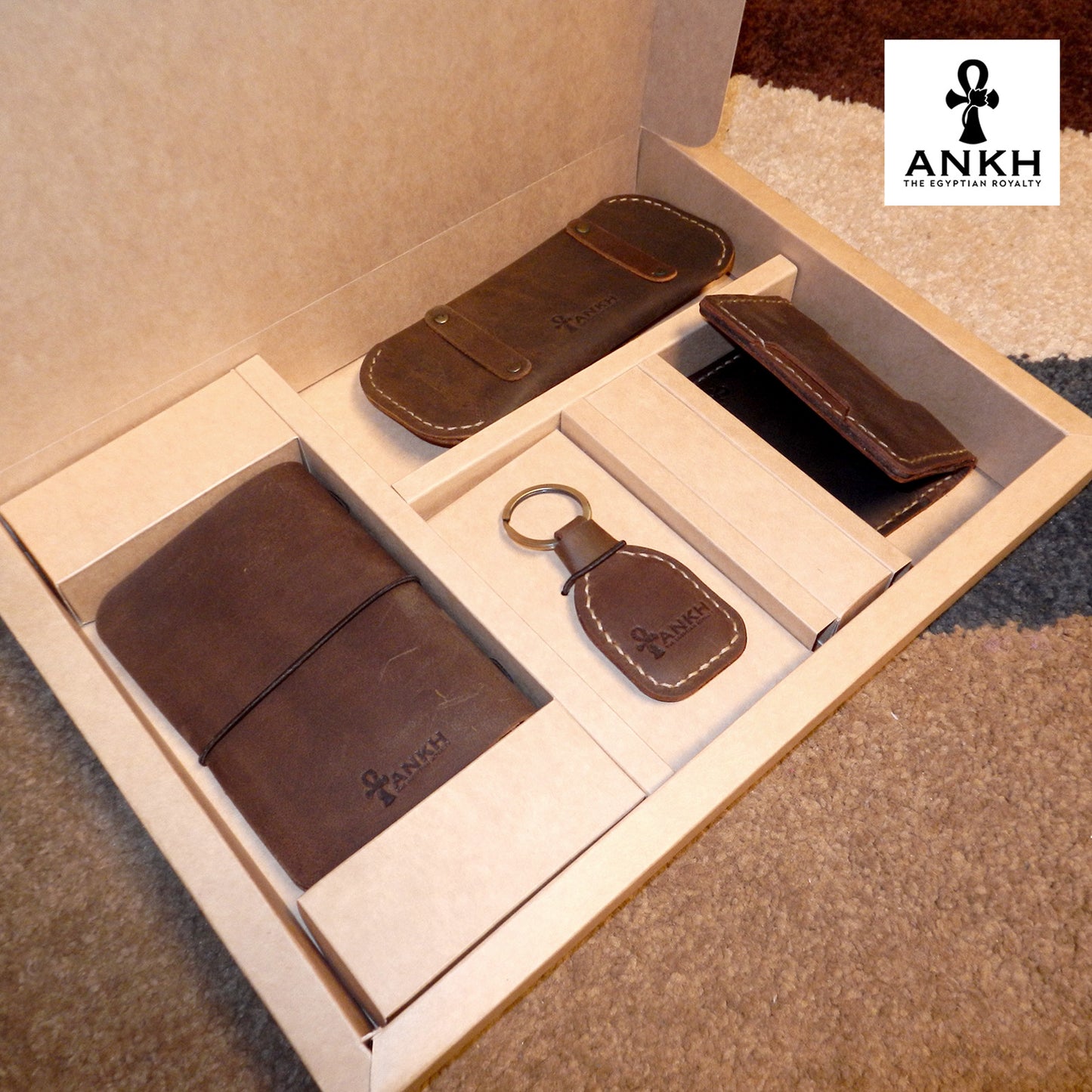 A white box containing four ANKH leather accessories: a passport cover with notebook, a key chain, a glasses cover, and a card holder, displayed on a textured surface. طقم هدية من الجلد الطبيعي الفاخر منقوش برمز العنخ المصري هدية من مصر