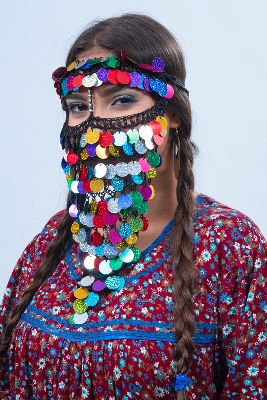 A woman wearing a Burqa (Veil) made of red, green, and gold beads that form geometric patterns and hang from a metal band around her forehead. She has dark eyes and red lips, and looks at the camera with a mysterious expression.