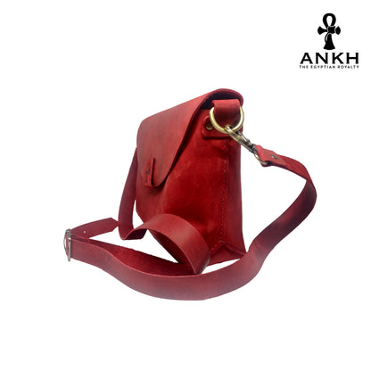 A genuine leather cross bag in red color with handcrafted brass accessories and multiple pockets, displayed on a white mannequin bust with a black logo of ANKH - The Egyptian Royalty.