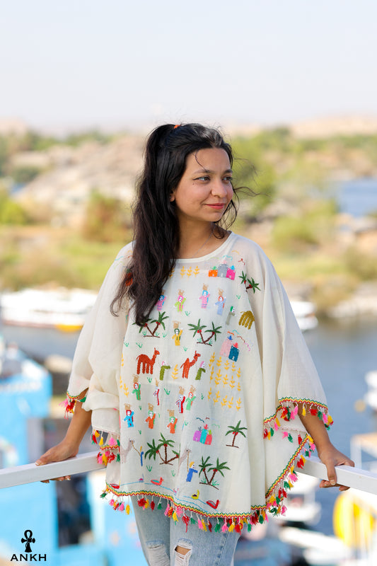 A woman wearing an off-white cotton blouse with bright colored embroidery inspired by Egyptian rural culture, standing against a railing with natural scenery in the background.