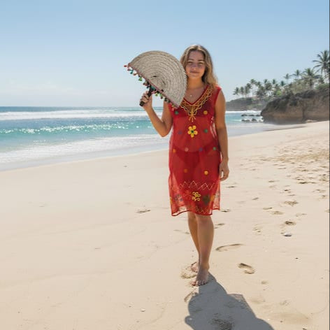 Woman in a red dress holding a straw hat on a beach with palm trees in the background