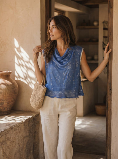 Woman in a blue sleeveless tally top and beige pants standing in a sunlit room with rustic decor.