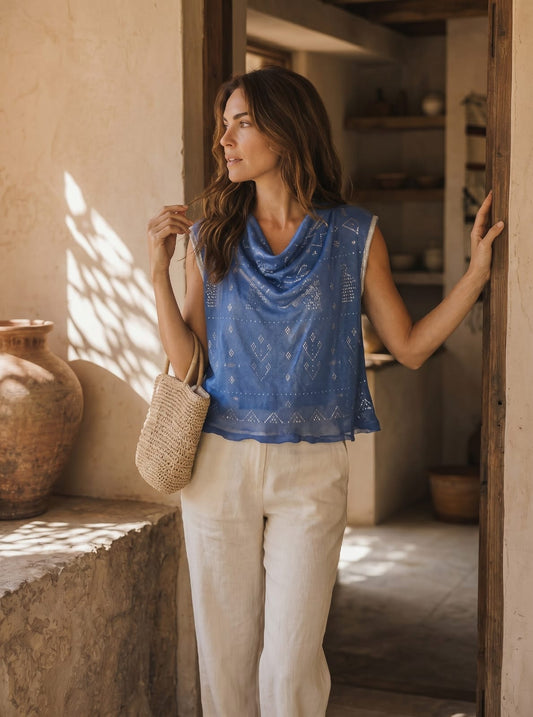 Woman in a blue sleeveless tally top and beige pants standing in a sunlit room with rustic decor.