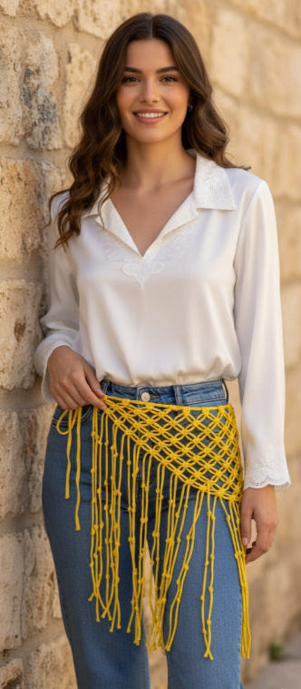 Woman wearing a white blouse and blue pants with a yellow fringe accessory against a stone wall.