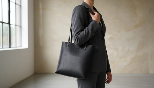 Person wearing a dark gray suit holding a black tote bag in a minimalistic room.