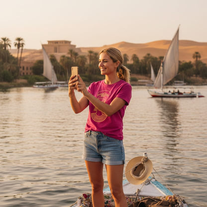 Woman taking a selfie on a boat with Felucca sailboats and ancient Egyptian architecture in the background.