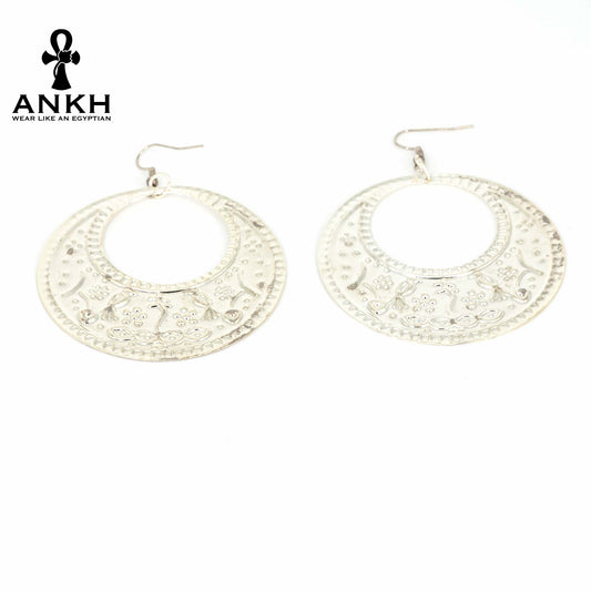 A pair of silver earrings with intricate engravings inspired by traditional Egyptian artistry, Egyptian accessory piece displayed on Egyptian woman model wearing traditional gallabia for women and heritage Egyptian Accessories, with a black logo of ANKH